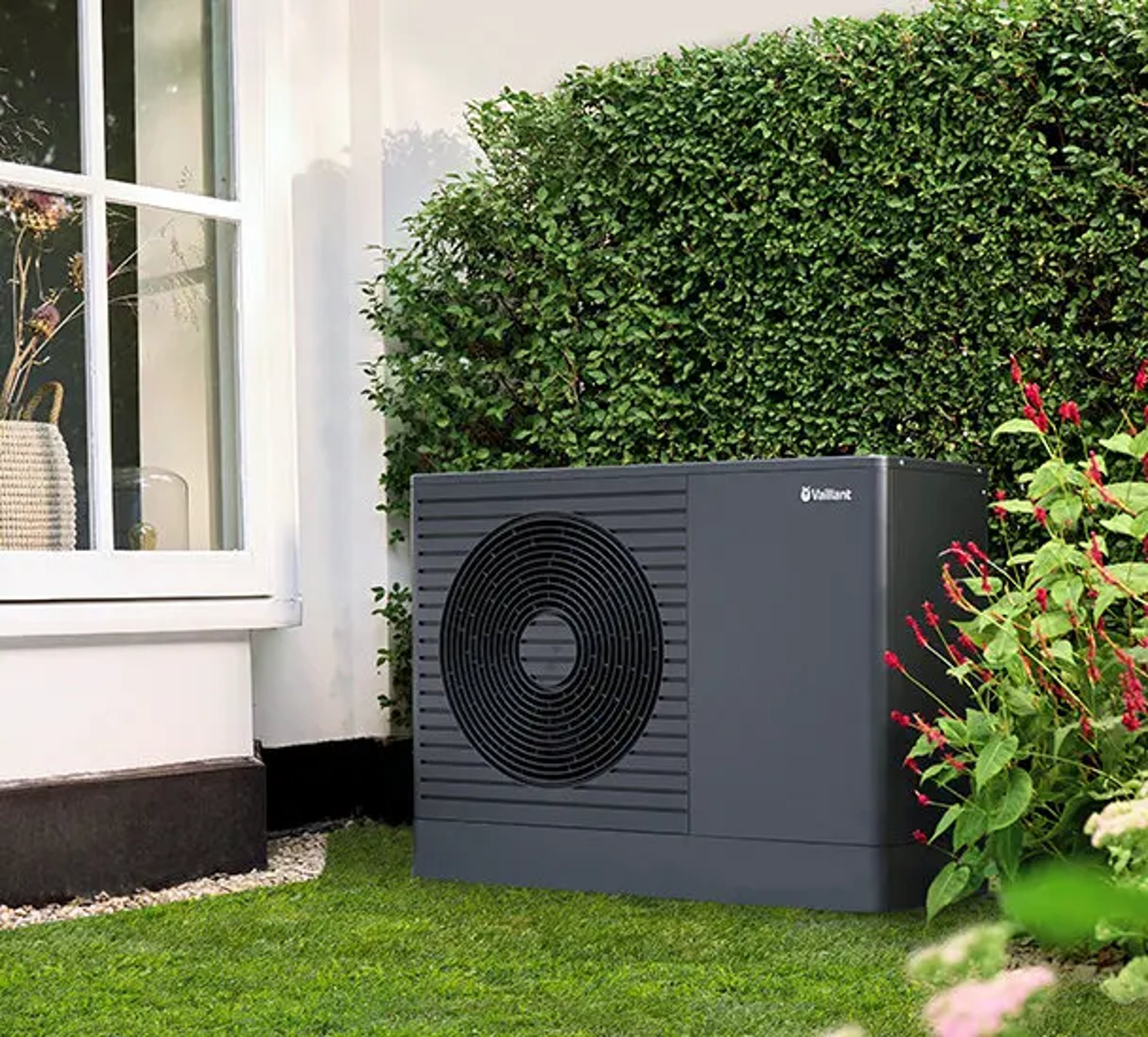 Dunkelgraue Vaillant aroTHERM plus Wärmepumpe im Garten neben einem Fenster und einer Hecke.